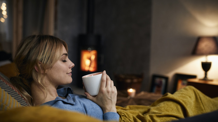 Eine Frau sitzt mit einer Decke auf dem Sofa, trinkt ein warmes Getränk, das Feuer brennt im Kamin und das Licht ist gedimmt.