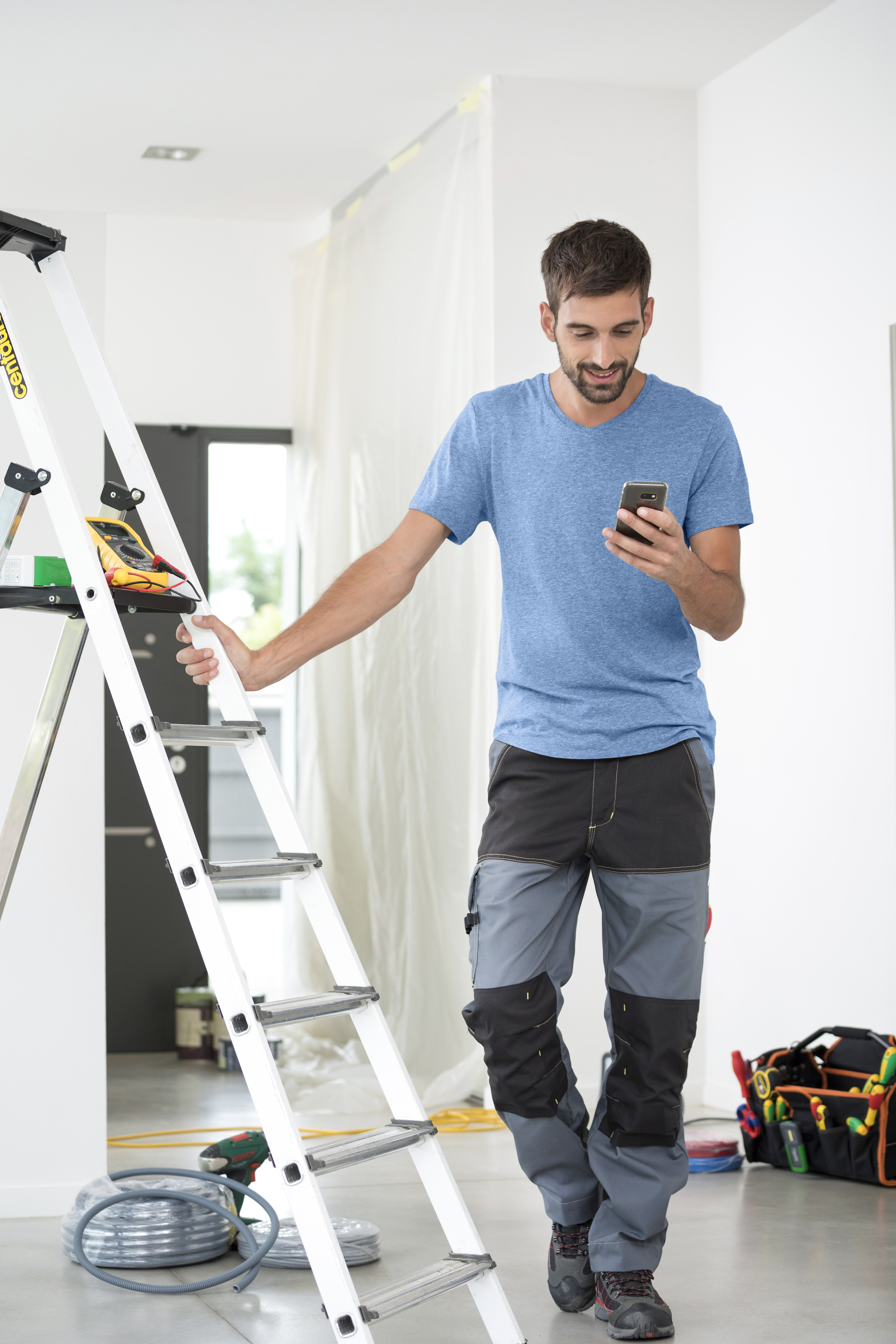 Elektriker mit Smartphone an einer Leiter lehnend.
